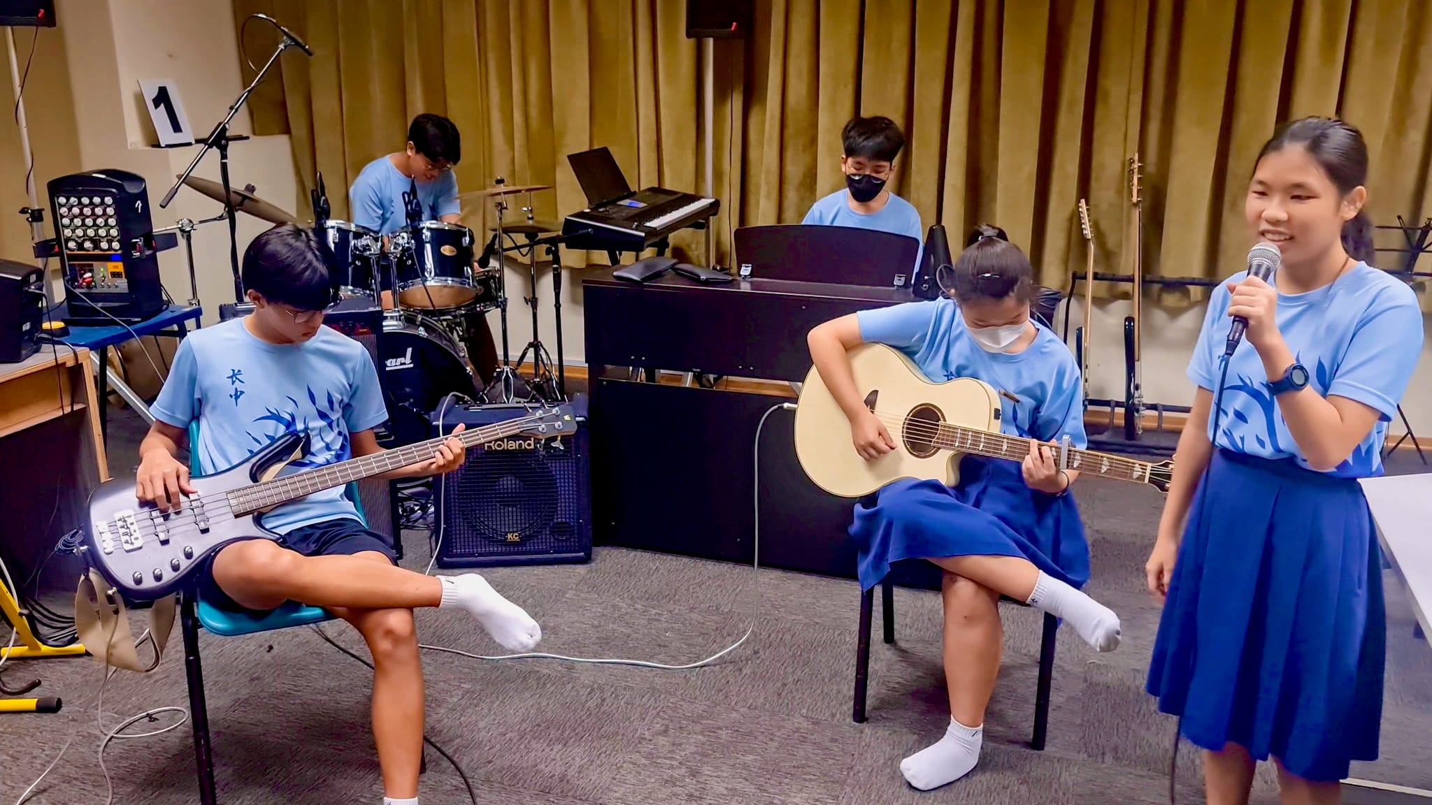 Five students in blue uniforms perform in a band with guitars, drums, keyboard, and a vocalist holding a microphone.